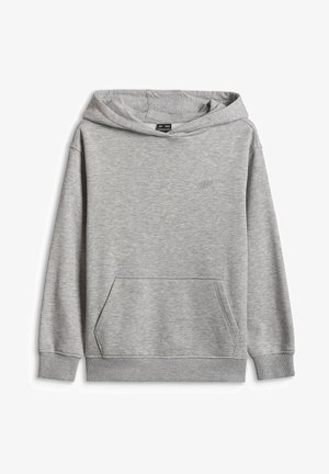 Enfant debout, face à l’objectif, portant un sweat à capuche gris clair, un pantalon gris ample et des baskets blanches sur un fond uni.