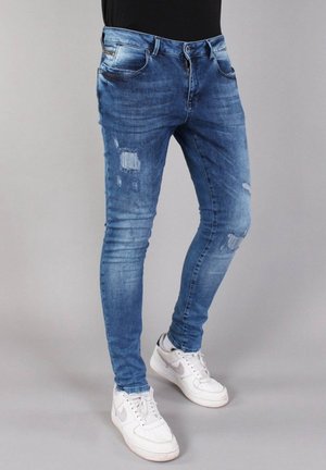 Person trägt schlank geschnittene, abgenutzte blaue Jeans und weiße Nike-Sneaker, steht vor einem einfarbigen hellgrauen Hintergrund.