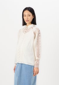 Witte, lichte blouse met ingewikkelde kantpatronen langs de mouwen en de knopenlijst; heeft een kraaghalslijn en een ontspannen pasvorm.