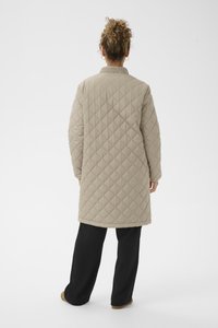 Beige quiltet lang jakke med ribbet krave, prydet med et diamantmønster og en afslappet pasform, vist fra ryggen sammen med sorte bukser.