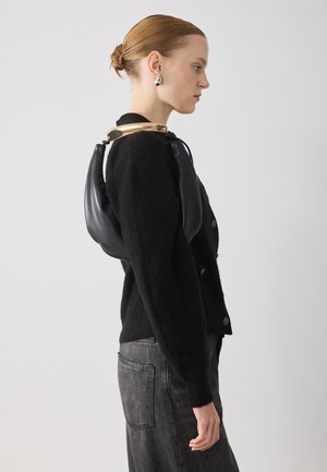 Borsa in pelle nera con design curvo, indossata a spalla. Presenta un accento metallico oro alla scollatura, abbinata a un maglione nero texture.