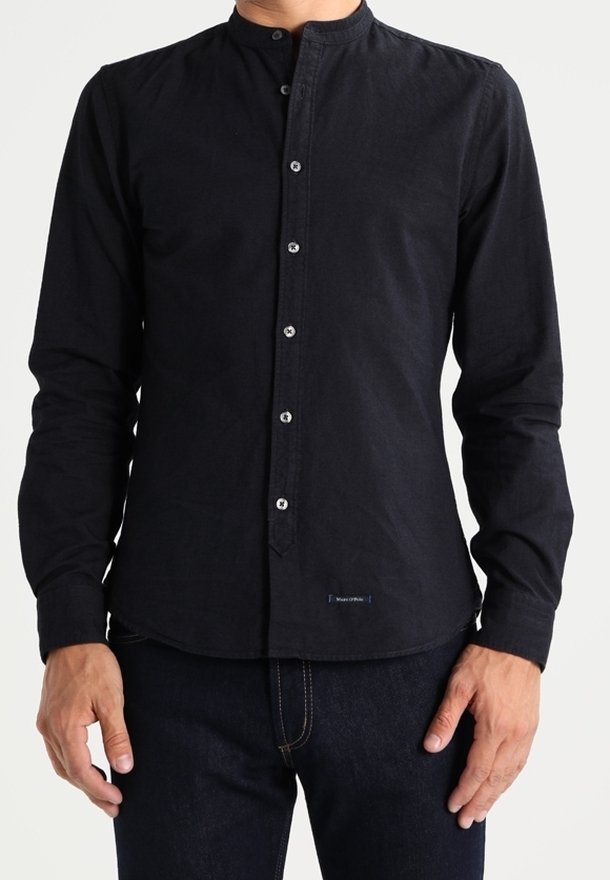 Camisa negra con botones, cuello mao, mangas largas y textura sutil. Presenta botones plateados y una pequeña etiqueta en el dobladillo.