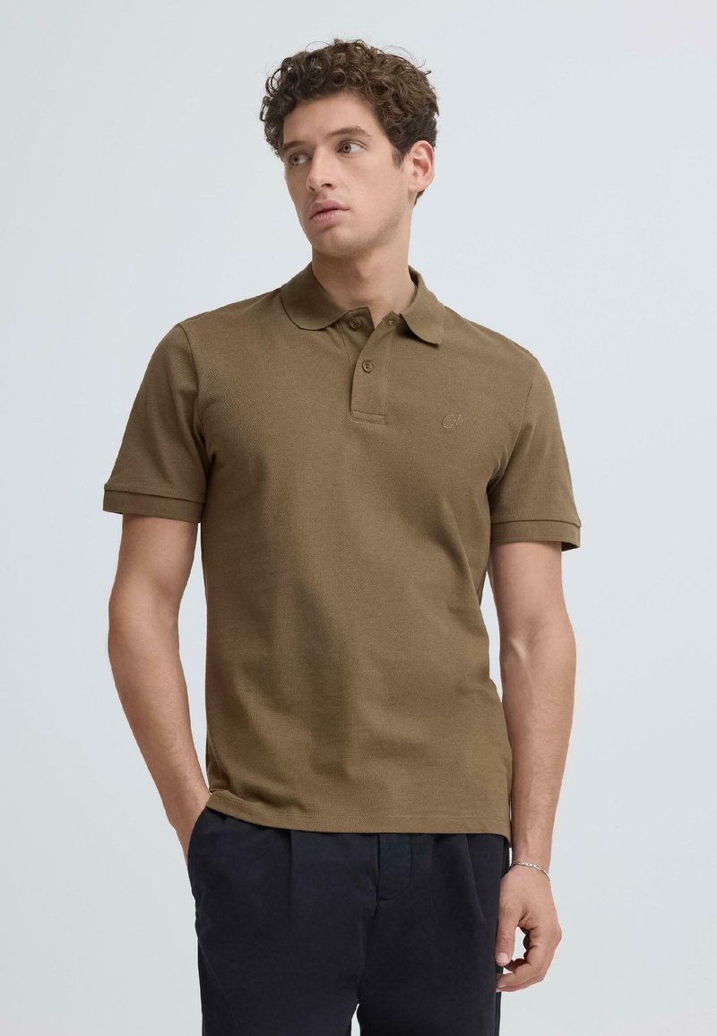 Jeune homme aux cheveux bouclés portant un polo marron à manches courtes et un pantalon sombre, debout avec les mains partiellement dans les poches.