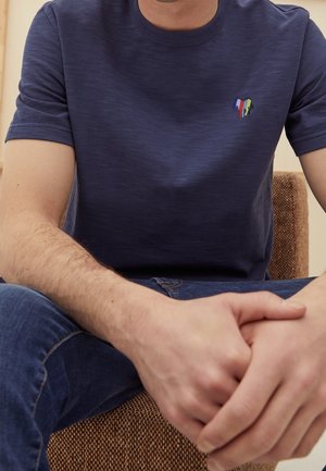 Homme portant un t-shirt bleu foncé avec un petit logo multicolore, assis les mains jointes sur les genoux, portant un jean bleu sur une chaise marron.