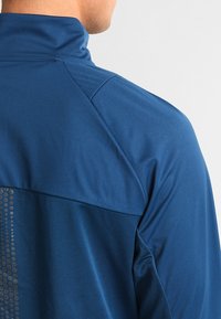 Uomo che indossa una giacca sportiva blu con colletto alto, mostrato di spalle con dettagli visibili della trama del tessuto e delle cuciture.