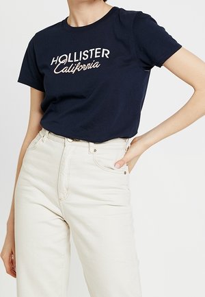 Vrouw draagt een marineblauw Hollister California T-shirt, in een high-waisted off-white spijkerbroek gestopt, één hand op haar heup.