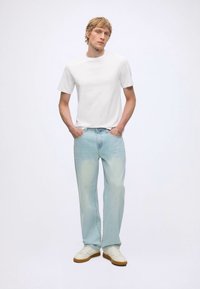 Junger Mann steht mit den Händen in den Taschen, trägt ein schlichtes weißes T-Shirt, hellblaue Jeans und weiße Sneaker vor weißem Hintergrund.