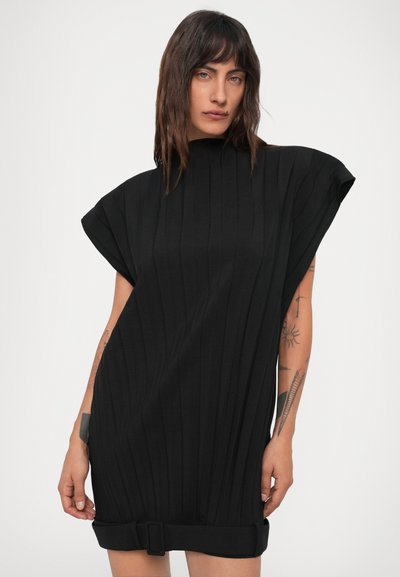 Pessoa com cabelo escuro até aos ombros, vestindo um vestido preto solto com textura de malha, com ombros largos e um detalhe de cinto na bainha, tatuagens visíveis nos braços.