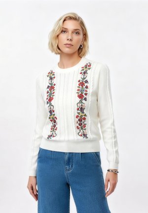 Femme aux cheveux blonds courts portant un pull blanc avec une broderie florale verticale et un jean bleu, debout les bras détendus.