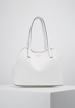 Hvid Guess håndtaske i læder med dobbelte skulderremme og guldtone logo, placeret på en hvid piedestal mod en lys grå baggrund.