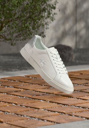 Baskets blanches Calvin Klein avec détail argenté au talon flottant au-dessus d'un bloc de bois et d'une surface de galets en extérieur.