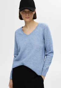 Svijetloplavi pulover s v-neck izrezom napravljen od mekanog materijala, s dugim rukavima i blago skraćenim rubom, uparen s crnom kapom.
