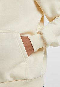 Crème-kleurige hoodie met een kangaroo-zak, ribgebreide manchetten en een soepele textuur, die een eenvoudig, functioneel ontwerp benadrukt.