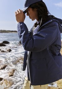 Giacca impermeabile blu navy con cappuccio, chiusura con zip e polsini elastici. Abbinata a una camicia azzurro chiaro e pantaloni beige, con un cappello coordinato.