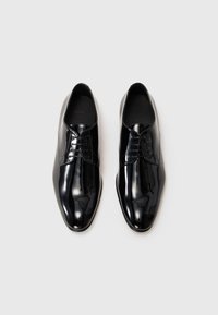 Zapatos de vestir de hombre de cuero negro pulido con cordones, vistos desde arriba sobre un fondo claro y liso.