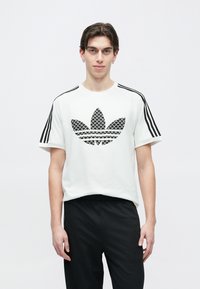 Fehér póló fekete mintás Adidas logóval, fekete csíkozott vállrésszel. Fekete, texturált nadrággal párosítva.