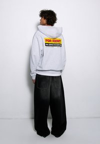 Szary hoodie z kapturem z dużą grafiką "DO WYNAJĘCIA" w czerwonym i żółtym na plecach, w połączeniu z oversizowymi czarnymi dżinsami.