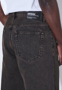 Tummansiniset farkut, joissa on hieman haalistunutta sävyä, takataskut ja musta vyötäröllä oleva etiketti valkoisella tekstillä.