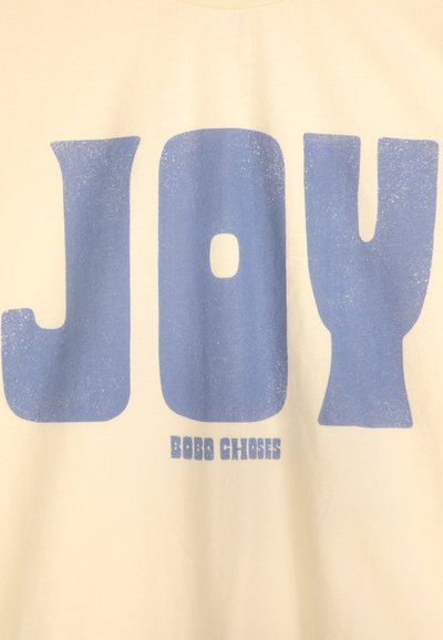 Cremefarvet t-shirt med store blå bogstaver, der staver "JOY" med en centreret hvid oval, og "BOBO CHOSES" i mindre blå tekst nedenfor.