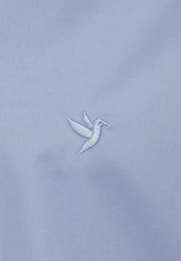 Una tela azul claro presenta un pequeño diseño bordado de un colibrí blanco, capturando los detalles de la forma y las alas del pájaro.