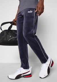 Σαλοπέτα ναυτικού βελουτέ παντελόνι με λευκές ραφές, διαθέτοντας λογότυπο. Συνδυασμένα με αθλητικά παπούτσια Puma, υφαντό ύφασμα και μια μαύρη τσάντα duffel.