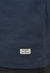 Marineblå bomulls-T-skjorte med en sydd etikett nederst med teksten "BLEND 1993 BYGGET I BLÅ." Slett tekstur, uformell design.