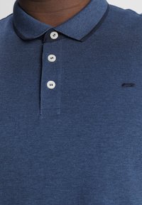 Marineblå polo skjorte med struktureret stof, tre-knaps placket og kontrastfarvet krave. Har et lille logo nær kanten.