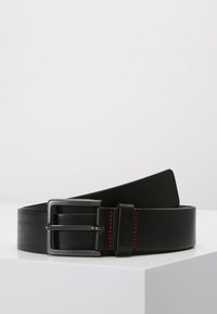 Curea din piele neagră cu o cataramă din metal argintiu. Prezintă cusături roșii de-a lungul marginilor. Textură plată, netedă, cu un capăt conic.