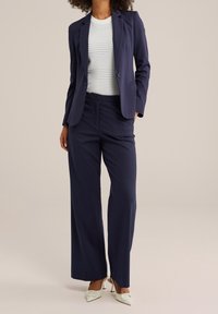 Woman wearing a navy blazer and wide-leg pants with a white perforated top and white pointed high heels, standing against a plain background.
