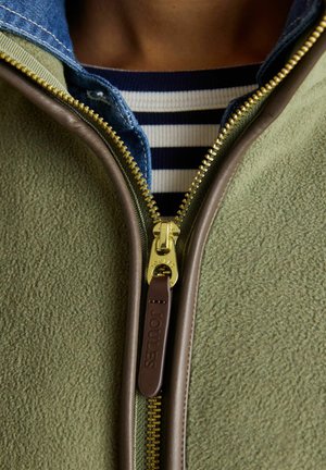 Detailný záber čiastočne rozopnutej olivovo zelenej flísovej bundy s koženým lemovaním cez rifľovú košeľu a námornícky modro-bielo pruhovaný top.