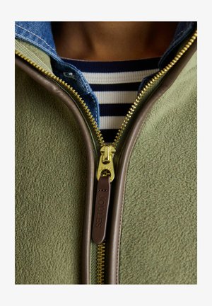 Zbliżenie na częściowo zasuniętą oliwkowozieloną polarową kurtkę z obszyciem ze skóry, noszoną na koszuli dżinsowej i granatowo-białym paskowanym topie.