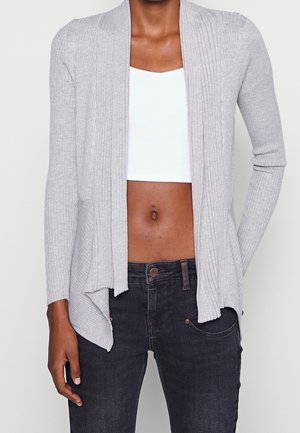 Frau trägt hellgrauen, gerippten Cardigan mit offenem Vorderteil, weißes Crop-Top und dunkle Jeans, steht mit einer Hand in der Tasche.