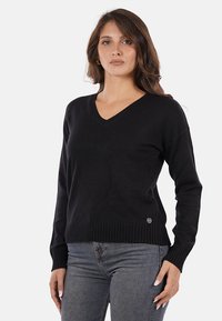 Maglione nero a V con polsini e orlo a costine, realizzato in tessuto morbido; esposto su una modella davanti a uno sfondo bianco semplice.
