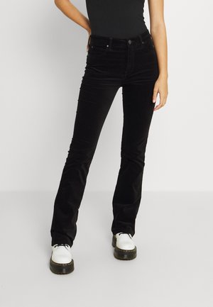 Vrouw draagt zwarte slim-fit broek en witte plateau schoenen, staat met één hand op haar heup tegen een effen achtergrond.