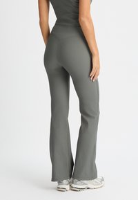 Pantaloni a zampa d'elefante in verde tenue, texture a coste, vita alta, abbinati a scarpe da ginnastica argentate. Vista posteriore focalizzata sul design e sulla vestibilità.