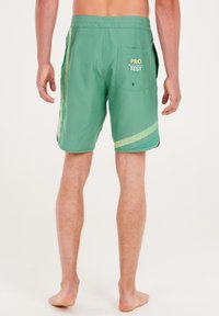 Groene zwemshorts met contrasterende lichtgroene strepen, een achterzak en "PRO TEST" in het geel gedrukt, gemaakt van lichtgewicht, sneldrogend materiaal.