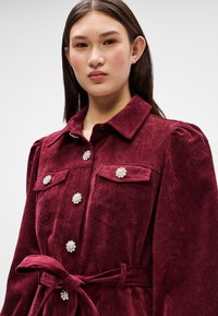 Burgundy kordbársony zakó, gallérral, virágmintás gombokkal, két mellzsebbel, és egy derékban megkötött azonos színű övvel.