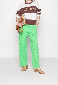Sudadera blanca y marrón con el texto "LONDON LEISURE CLUB", pantalones verdes de pierna ancha, sandalias de tacón marrones con tiras, sosteniendo un bolso tejido en tono marrón.