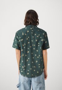 Chemise verte à manches courtes avec un petit motif de palmier, col boutonné et tissu doux. Comprend un ourlet arrondi et une coupe décontractée.