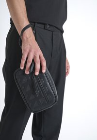 Bolso de muñeca de cuero negro con una superficie texturizada que presenta patrones geométricos y un cierre de cremallera, sostenido por una mano cerca de unos pantalones negros.