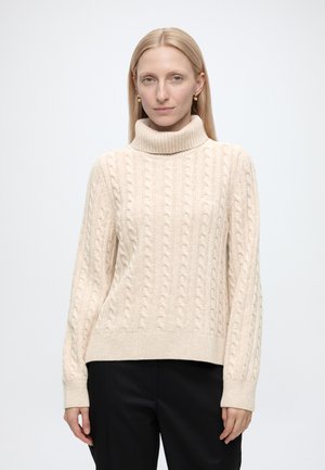 Frau mit glattem blondem Haar, die einen cremefarbenen Zopfmuster-Rollkragenpullover und schwarze Hose vor einem schlichten hellen Hintergrund trägt.