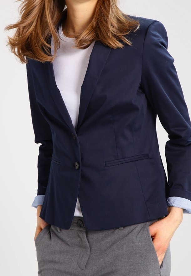 Femme portant un blazer bleu marine sur une chemise blanche aux manches retroussées et un pantalon gris, les mains dans les poches, devant un fond uni.