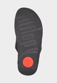 Svart gummisula på sandal med präglad "fitflop" text och en röd cirkulär detalj, med en texturerad yta och formad kontur.