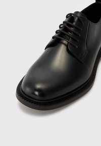 Black leather dress shoe with a smooth finish, round toe, and black laces. Features a textured black rubber sole and stitched accents.