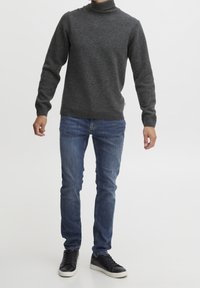 Pull à col roulé gris en tricot texturé, associé à un jean bleu slim. Le mannequin porte des baskets noires, debout devant un fond uni.
