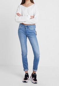 Weißer, verkürzter Pullover mit V-Ausschnitt, hellblaue Jeans mit aufgerollten Säumen und schwarze Sneakers mit pinken Akzenten.