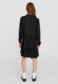 Woman with long brown hair wearing a long-sleeve black tiered dress and white ankle boots, standing facing away on a plain background.