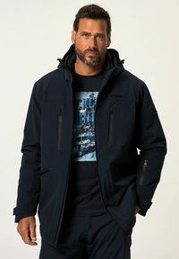 Marineblaue, wasserdichte Jacke mit Reißverschlusstaschen, Kapuze und taillierter Passform, kombiniert mit einem dunklen Grafik-T-Shirt mit einem Bergmotiv.