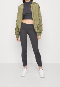 Olivgrön bomberjacka med samlade ärmar, svart kort topp och mörka leggings. Vita sneakers fulländar den sportiga looken.