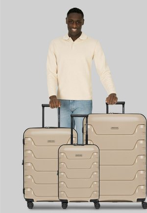 Homme portant une chemise beige à manches longues et un jean, debout derrière trois valises rigides beiges avec poignées extensibles sur fond gris.